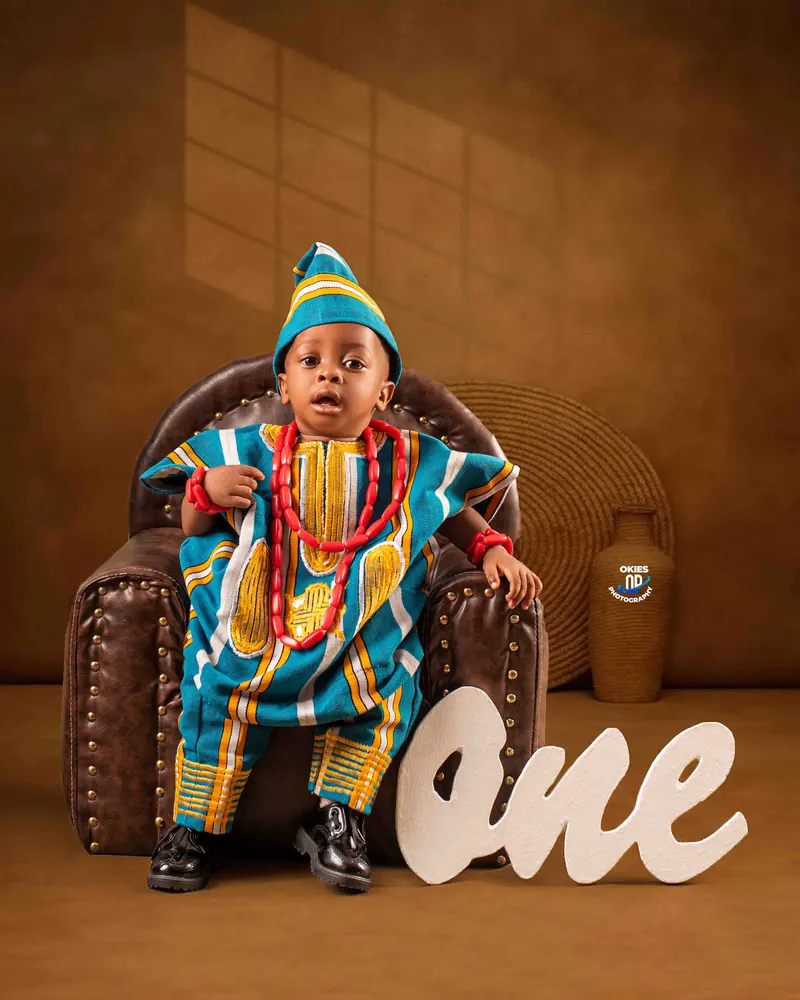A little boy in a bright aso-oke agbada and cap sits in a leather armchair beside a "One" sign for his first birthday