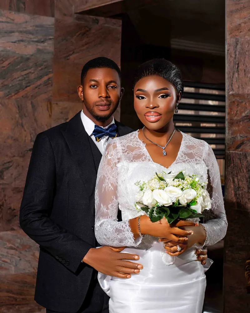 A groom in a dark suit and a bride in white lace holding white roses