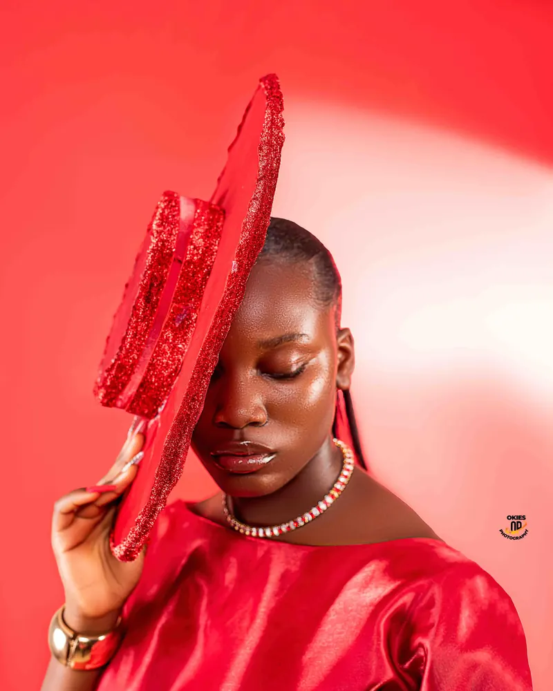 A woman in red tilts a wide red hat over one eye against a red backdrop