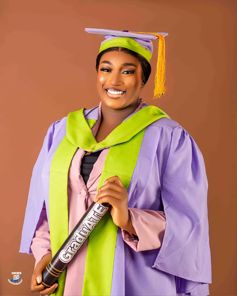 A smiling graduate in a lilac and lime gown and cap holds her certificate tube