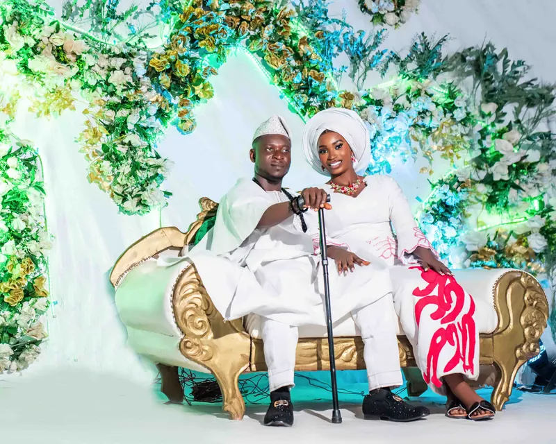 A couple in white traditional outfits sit on a gold sofa under a flower arch