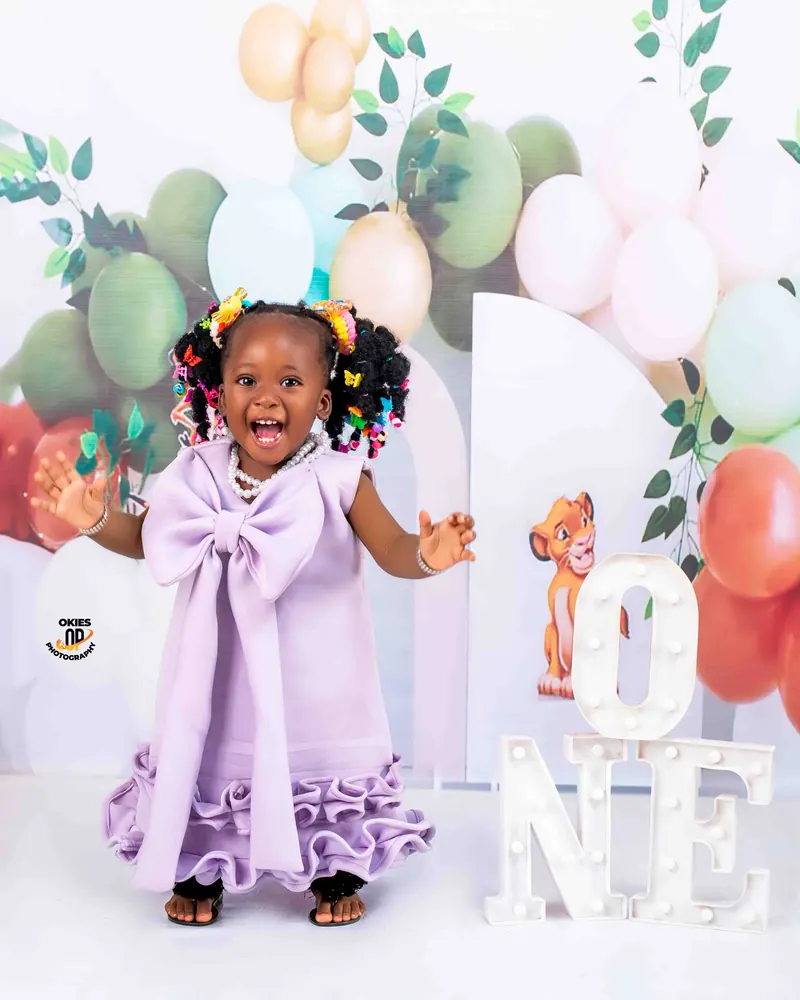 A toddler in a lilac dress with a big bow laughs beside "One" letters and balloons